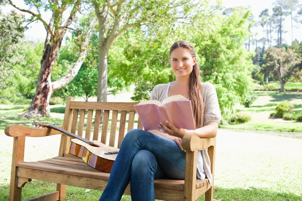 Χαμογελαστή γυναίκα με ένα βιβλίο και μια κιθάρα που κάθεται σε ένα παγκάκι — Φωτογραφία Αρχείου