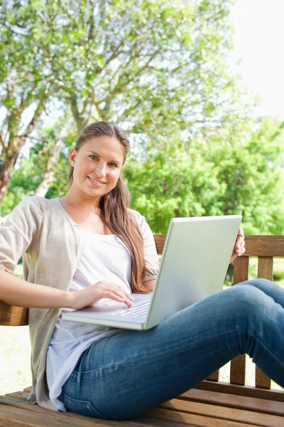 Uśmiechający się z laptopa, siedząc na ławce w parku — Zdjęcie stockowe
