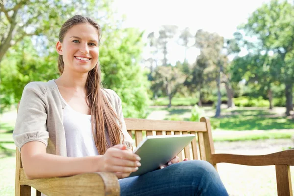微笑着坐在她的平板电脑与公园长椅上的女人 — 图库照片