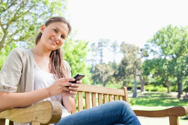 Mulher sorridente escrevendo uma mensagem de texto em um banco de parque — Fotografia de Stock