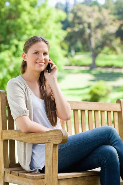 Sorrindo mulher sentada com seu telefone celular em um banco de parque — Fotografia de Stock