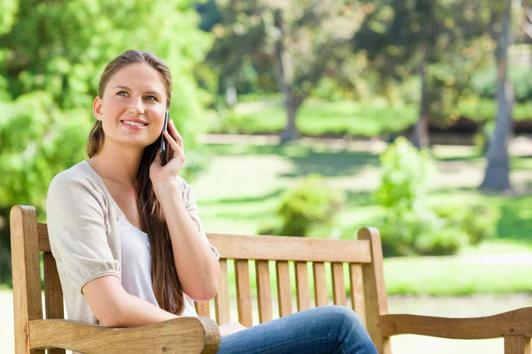 Usmívající se žena na její mobil, zatímco sedí na lavičce v parku — Stock fotografie
