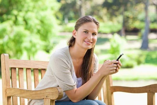 Улыбающаяся женщина, сидящая на скамейке в парке со своим телефоном — стоковое фото