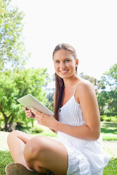 Vista lateral de una mujer sonriente sentada en el césped con una tableta c —  Fotos de Stock