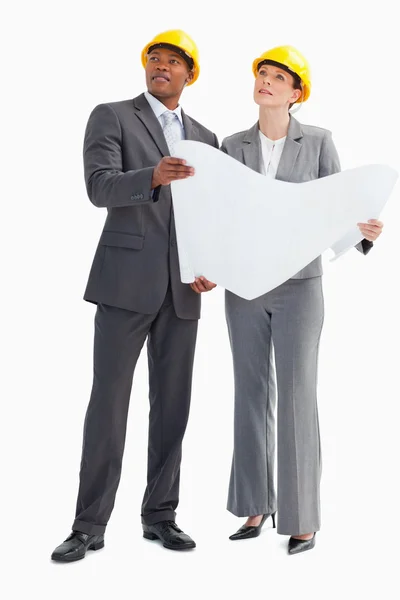 Businessman and woman wearing hard hats holding paper looking up — Stock Photo, Image