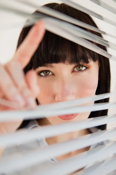 A woman looking through some blinds into the camera — Stock Photo, Image
