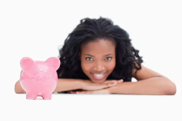 Ein lächelndes Mädchen, das seinen Kopf auf den Händen mit einem Sparschwein ruht — Stockfoto