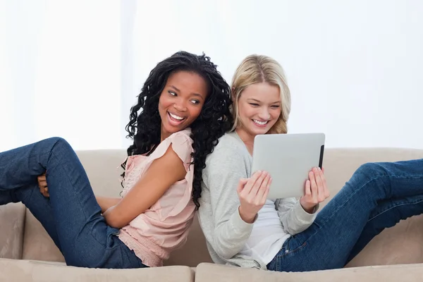 Dos mujeres sentadas espalda con espalda en un sofá mirando una tableta co —  Fotos de Stock