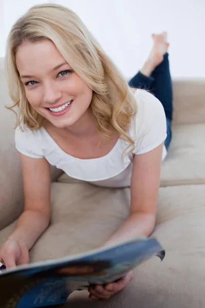 Une femme souriante est allongée sur un canapé et tient un magazine — Photo