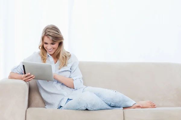 Uma mulher sorrindo segurando um tablet está sentada em um sofá — Fotografia de Stock