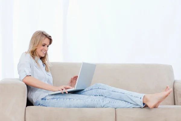 Una mujer está acostada en un sofá escribiendo en su portátil —  Fotos de Stock