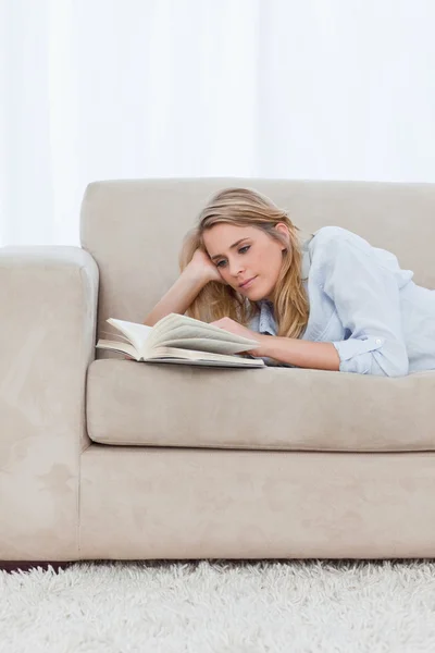 Una mujer acostada en un sofá apoyando su cabeza sobre su mano está leyendo — Foto de Stock