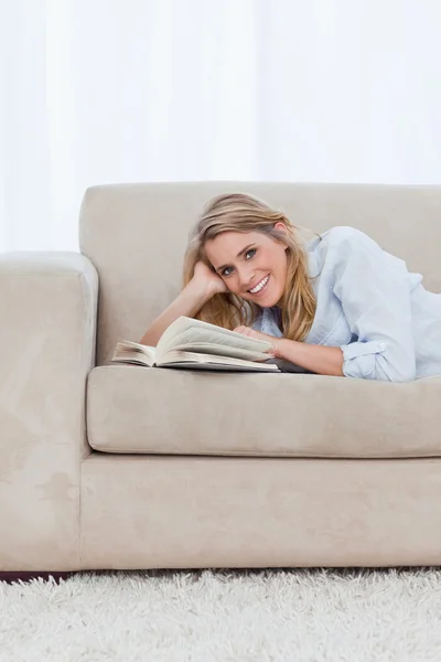 A woman looking at the camera is lying on a couch with a book — Stock Photo, Image