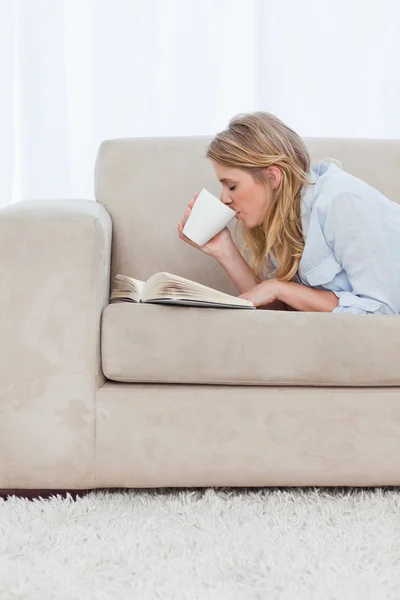 Bir kitapla kanepede yatan bir kadın kahve içiyor — Stok fotoğraf
