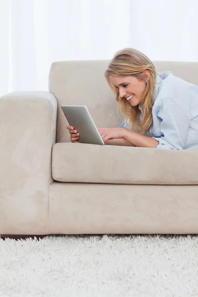 Uma mulher sorrindo está deitada em um sofá usando um tablet — Fotografia de Stock