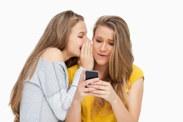 Estudante sussurrando para sua amiga que está enviando mensagens em seu telefone — Fotografia de Stock