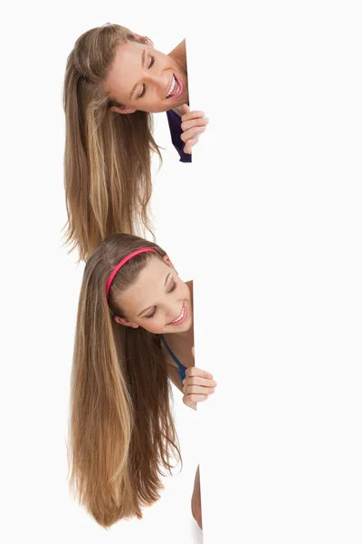 Close-up de dois estudantes de cabelos longos sorridentes atrás de um sinal em branco — Fotografia de Stock