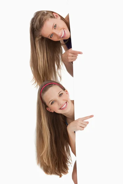 Retrato de dos estudiantes de pelo largo señalando detrás de un signo en blanco —  Fotos de Stock