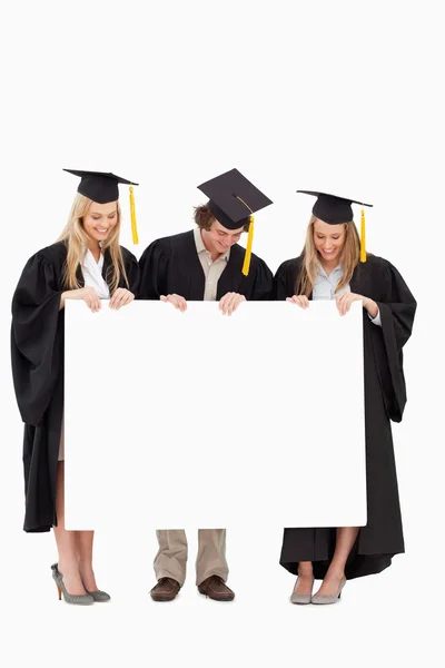 Três estudantes sorrindo em roupão de graduação segurando um sinal em branco — Fotografia de Stock