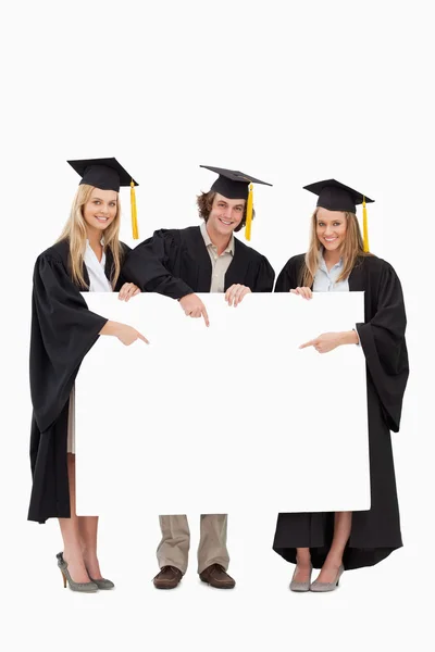 三名学生在毕业袍持有并指向空白 sig — 图库照片