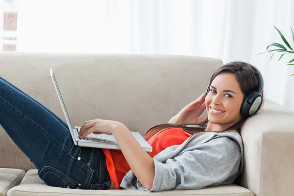A woman looking at the camera while she uses a laptop and headph — Stock Photo, Image