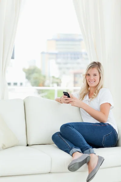 En kvinna som sitter i sidled på soffan som hon använder sin telefon och — Stockfoto