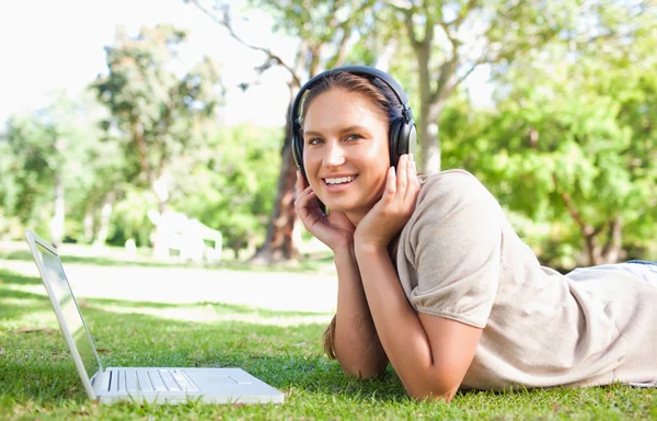 有一台笔记本电脑和耳机躺在草坪上的女人 免版税图库图片