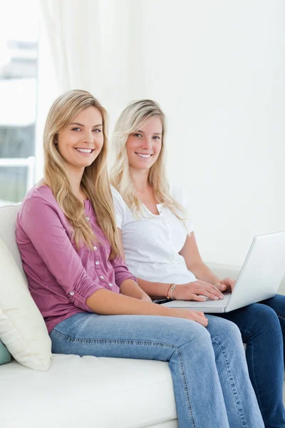 Two sisters sit on the couch with a laptop as they smile at the — Stock Photo, Image