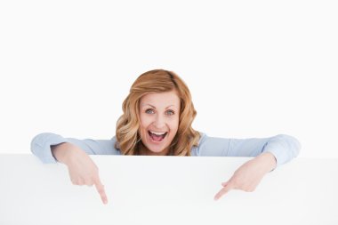 Happy blond-haired woman standing behind an empty white board clipart