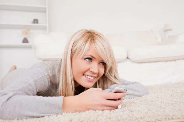 Pretty blond woman playing with cell phone lying down on a carpe — Stock Photo, Image