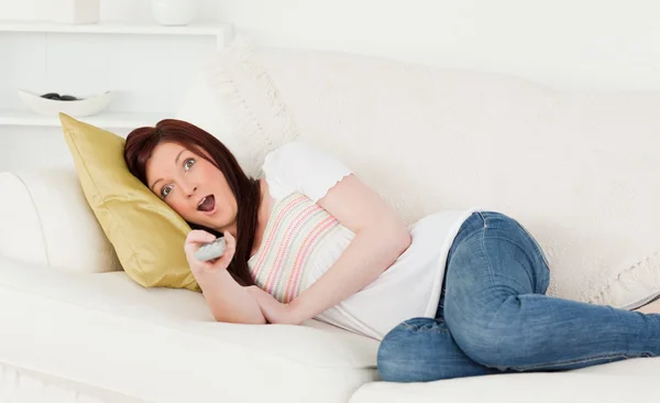 Attractive red-haired woman watching tv while lying on a sofa — Stockfoto