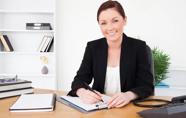 Beautiful red-haired female in suit writing on a notepad and pos — Stock Photo, Image