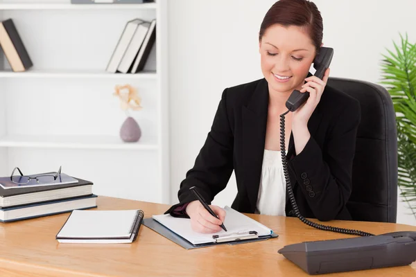 Beautiful red-haired woman in suit writing on a notepad and phon — Stock Photo, Image
