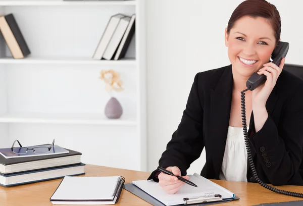 Pretty red-haired woman in suit writing on a notepad and phoning — Stock Photo, Image