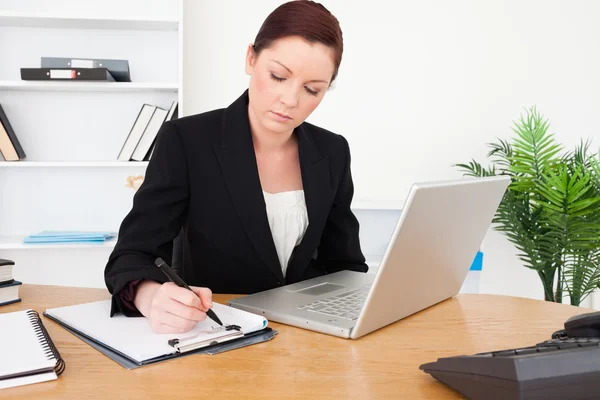 Young beautiful red-haired female in suit typing on her laptop a Stockafbeelding