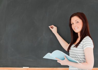 Young woman about to write on a blackboard looking at the camera clipart