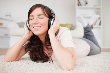 Smiling brunette woman using headphones while lying on a carpet clipart