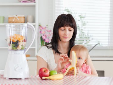 Cute brunette woman pealing a banana while holding her baby on h clipart