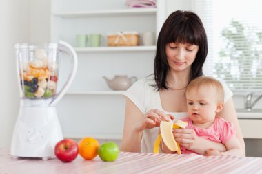 Gorgeous brunette woman pealing a banana while holding her baby clipart