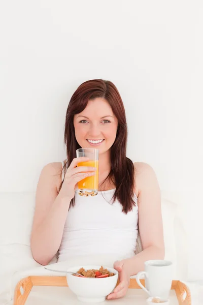 Bella femmina dai capelli rossi che beve un bicchiere di succo d'arancia mentre — Foto Stock