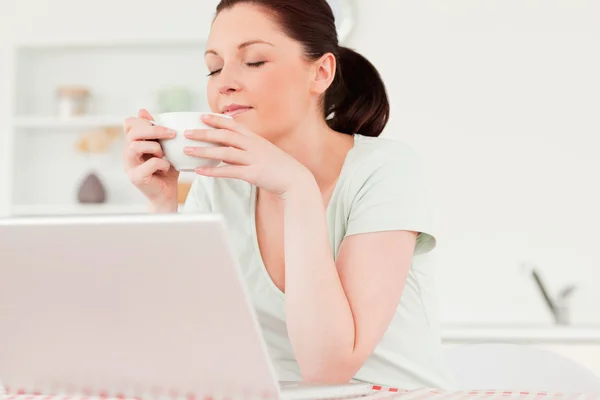 Gut aussehende Frau entspannt sich auf ihrem Laptop, während sie eine Tasse trinkt — Stockfoto