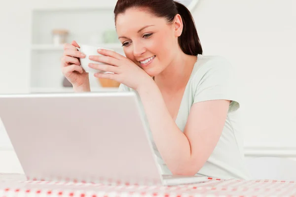 Encantadora mujer relajándose en su computadora portátil mientras bebe una taza de té —  Fotos de Stock