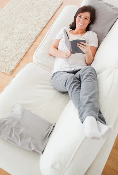 Junge hübsche Frau liest ein Buch, während sie auf einem Sofa liegt — Stockfoto