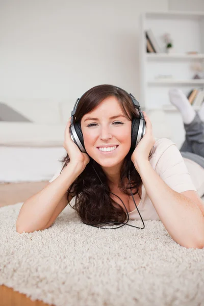 Mulher muito sorridente usando fones de ouvido enquanto deitada em um tapete — Fotografia de Stock