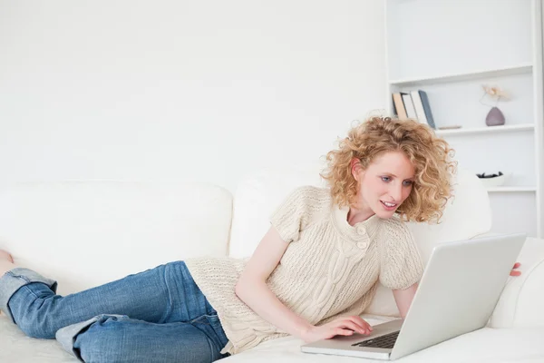Ładny blond kobieta relaksując się przy jej laptop leżąc na tak — Zdjęcie stockowe