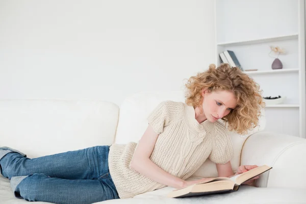 Schöne blonde Frau liest ein Buch, während sie auf einem Sofa liegt — Stockfoto