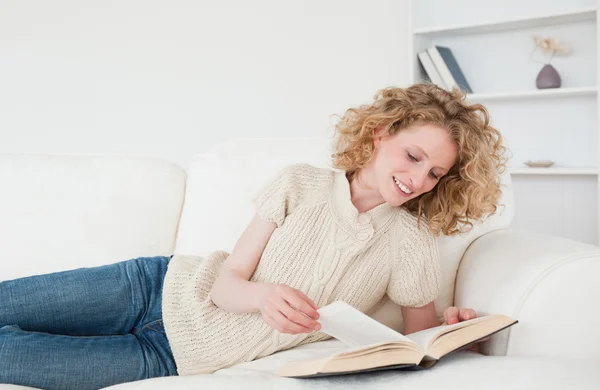 Attraktive blonde Frau liest ein Buch, während sie auf einem Sofa liegt — Stockfoto