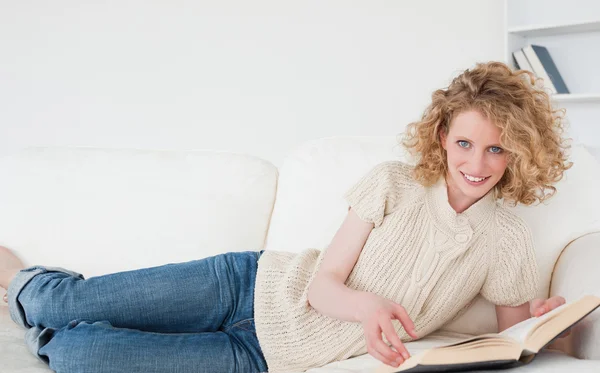 Hübsche blonde Frau liest ein Buch, während sie auf einem Sofa liegt — Stockfoto