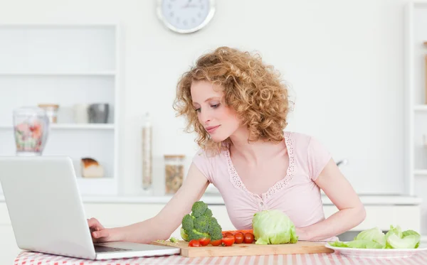 Mujer rubia bonita que se relaja con su computadora portátil mientras cocina un poco —  Fotos de Stock