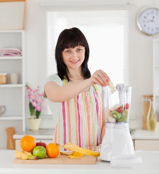 Schöne brünette Frau legt Gemüse in einen Mixer, während sta — Stockfoto